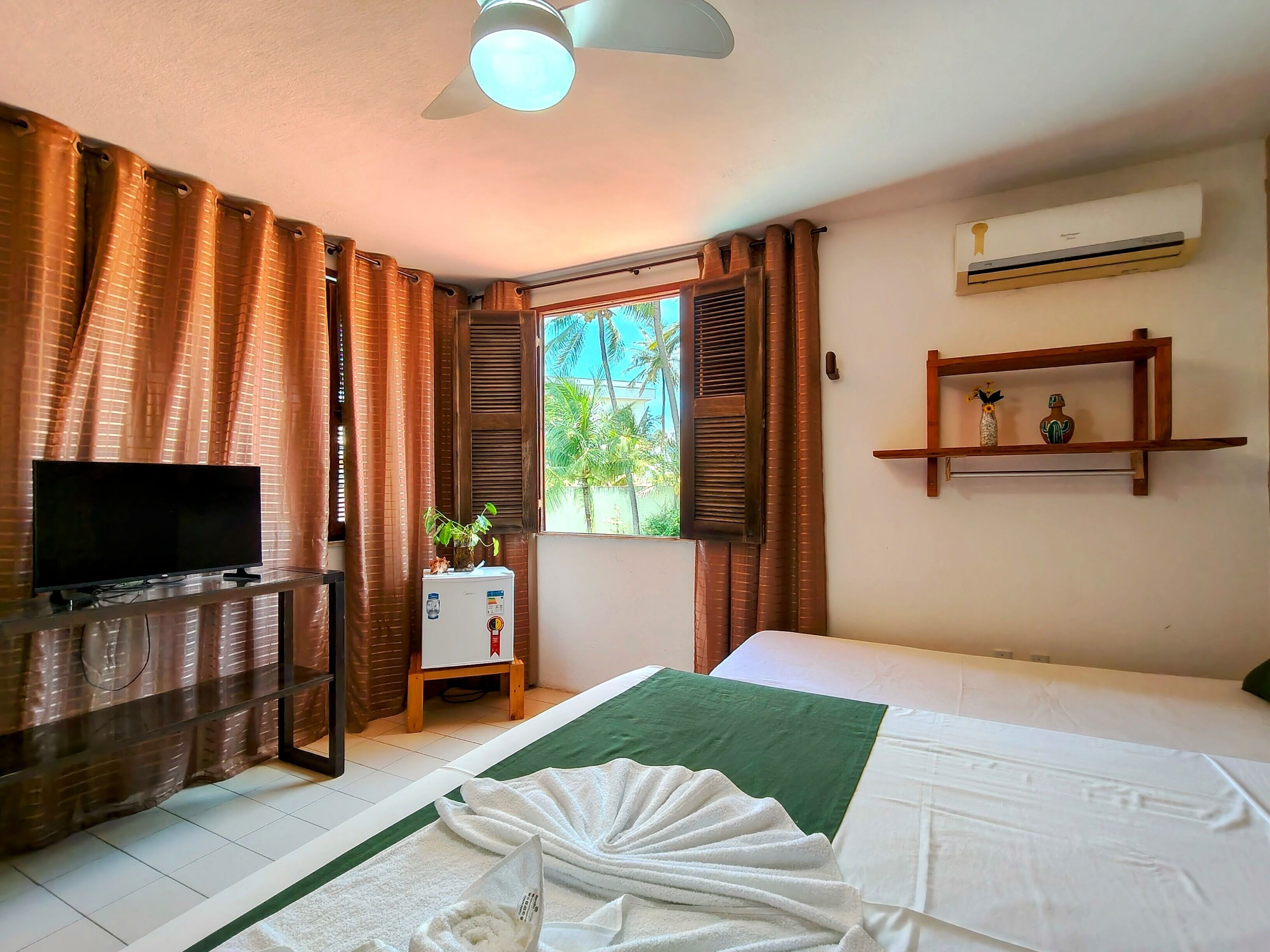 A hotel room with a bed, a TV, an air conditioner, a window with shutters, and a small table with a plant.