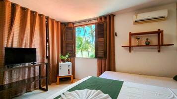 A hotel room with a bed, a TV, an air conditioner, a window with shutters, and a small table with a plant.