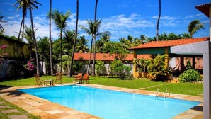 Une piscine dotée d’une terrasse carrelée, entourée de palmiers et bordée d’un bâtiment au toit rouge.
