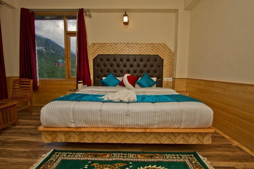 Quarto com uma cama grande, cabeceira de madeira, vista para montanhas pela janela e tapete verde com padrão.