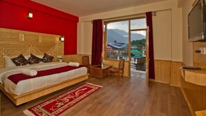 A hotel room with a large bed, wooden headboard, a small table, and a view of mountains and buildings through a glass door. - Mountain Green Villa (Manali)
