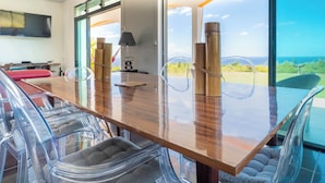 Uma sala de jantar moderna com mesa de madeira, cadeiras de plástico transparente e grandes janelas com vista para o mar.