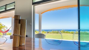 Uma mesa de madeira com tampo de vidro, uma rede e vista para o mar através de grandes janelas.