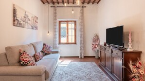 A living room with a sofa, a flat-screen TV, a wooden cabinet, a window with curtains, and a painting on the wall. - Casa Ilaria Vicopisano (Vicopisano)