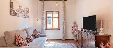 Ein Wohnzimmer mit einer Couch, einem Flachbildfernseher, einem Holzschrank, einem Fenster mit Vorhängen und einem Bild an der Wand.
