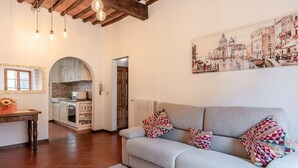 A living room with a sofa, patterned pillows, a painting of a cityscape, and a kitchen in the background. - Casa Ilaria Vicopisano (Vicopisano)