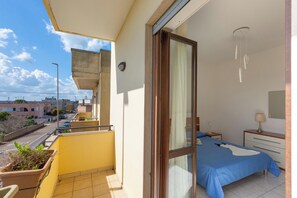 Un balcon avec un pot de fleurs, un lit recouvert d’une courtepointe bleue, une table de chevet avec une lampe, et une vue sur la rue et les immeubles.