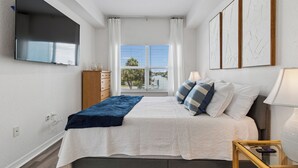 Une chambre à coucher avec un lit, un ventilateur de plafond, une télévision, une fenêtre avec des rideaux et des œuvres d’art accrochées au mur.