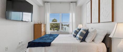 A bedroom with a bed, a ceiling fan, a TV, a window with curtains, and artwork on the wall.