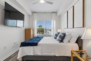 A bedroom with a bed, a ceiling fan, a TV, a window with curtains, and artwork on the wall. - Madeira Beach Escape  Pool + Water Views (Madeira Beach)