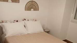 Una cama con colcha color beige, almohadas blancas y dos toallas blancas a un lado. Hay una mesita de noche con una lámpara.