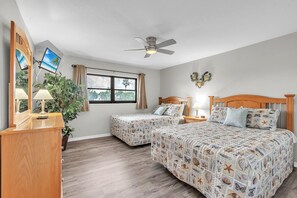 Ein Schlafzimmer mit zwei Betten, einem Deckenventilator, einem großen Fenster mit Vorhängen, einer Holzkommode und einem Spiegel.
