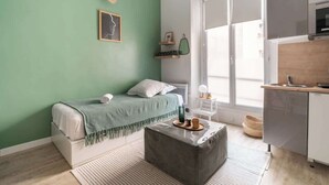 A modern bedroom with a bed, a bedside table, a grey ottoman, a window, and a kitchen area. - Studio Solo - Opera Galeries Fayette - 1 Pax (Paris)