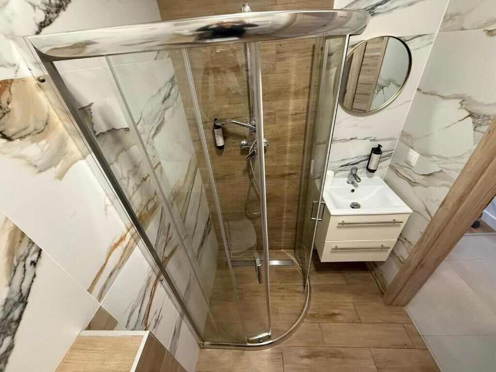 A glass-enclosed shower with a wooden floor, a white sink, and a marble-patterned wall.