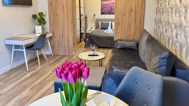 A modern living room with a sofa, a dining table with a vase of tulips, a TV mounted on the wall, and a sliding door leading to a bedroom. - Pokoje Balticus Mrzeżyno (Mrzeżyno)
