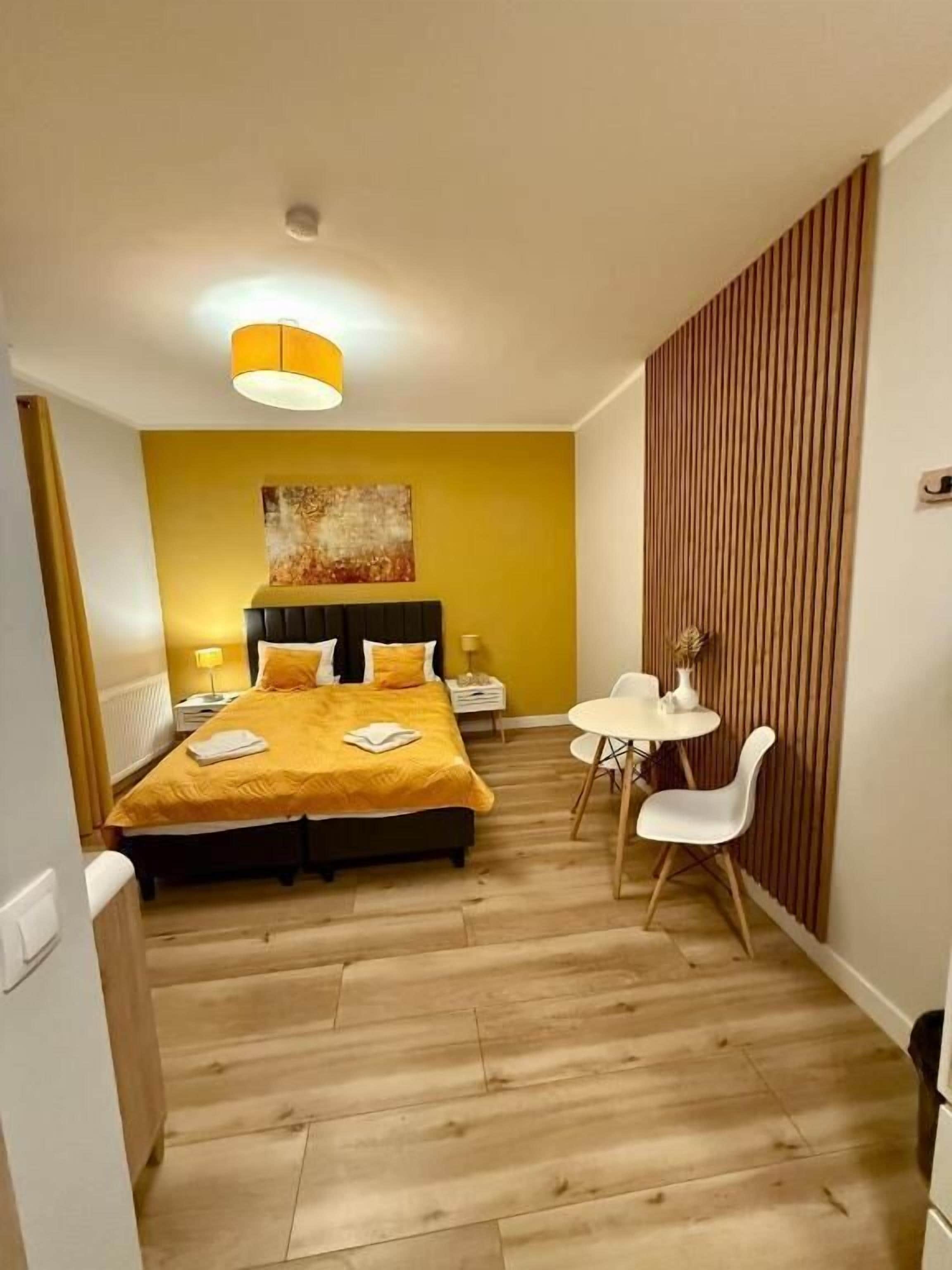 Une chambre d’hôtel comprenant un lit, des tables de chevet, un bureau avec des chaises, un plancher en bois et un mur d’accent jaune.
