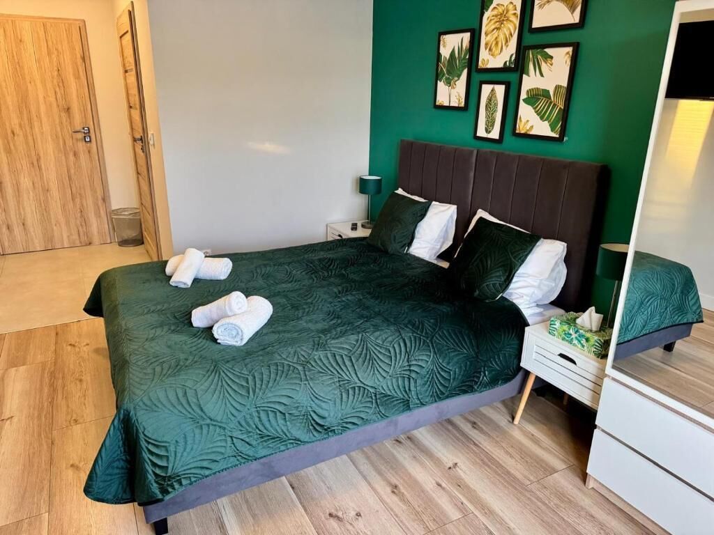 A neatly made bed with a dark green bedspread, two pillows, and rolled towels on a wooden floor.
