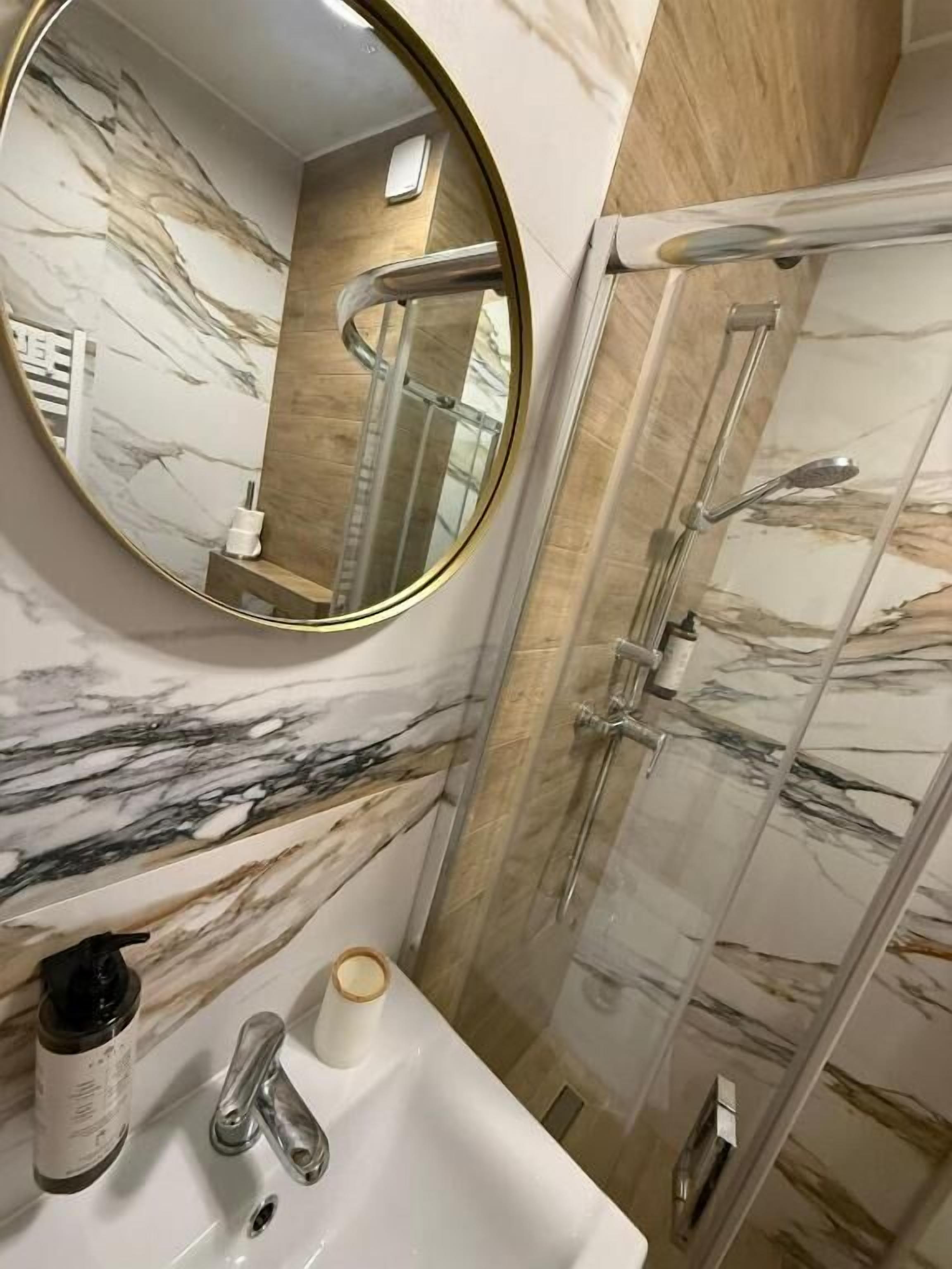 A bathroom with a marble wall, a round mirror, a sink with a faucet, and a glass shower enclosure.