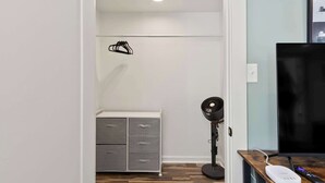 Una habitación con un armario blanco, un ventilador de pie, un televisor y una estantería en la pared.