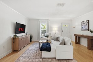 Ein modernes Wohnzimmer mit Flachbildfernseher, weißen Sofas, einem Couchtisch, einem gemusterten Teppich und einer Holzkommode.