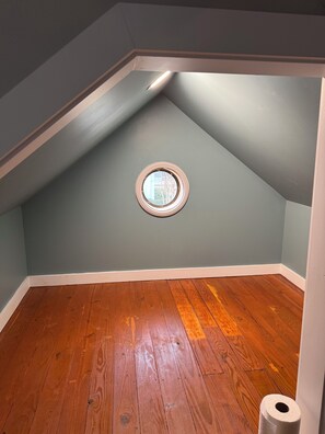 A room with a sloped ceiling, a small circular window, wooden flooring, and a white baseboard.