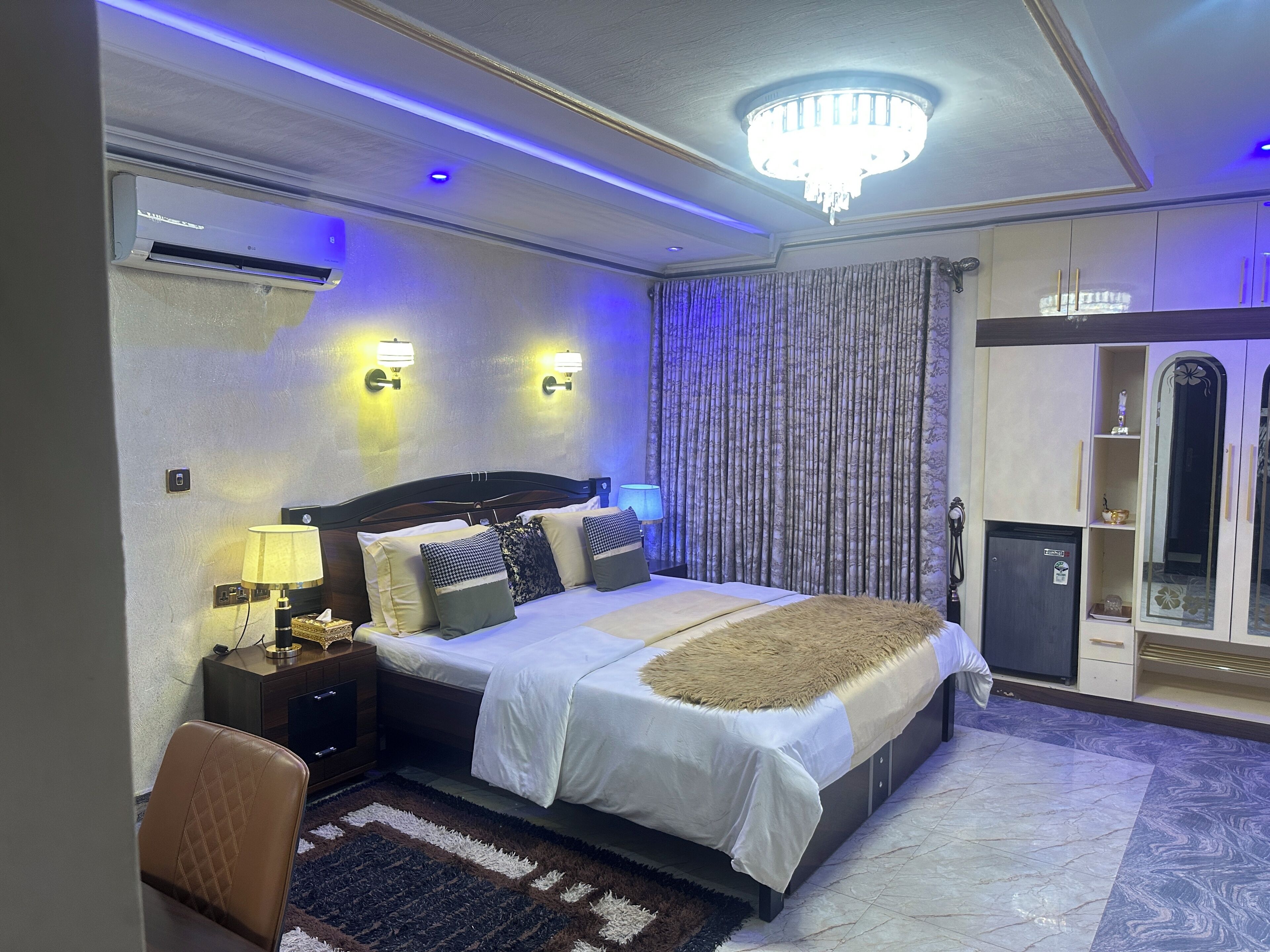 A hotel room with a bed, bedside lamps, a chandelier, a wardrobe, and a minibar.