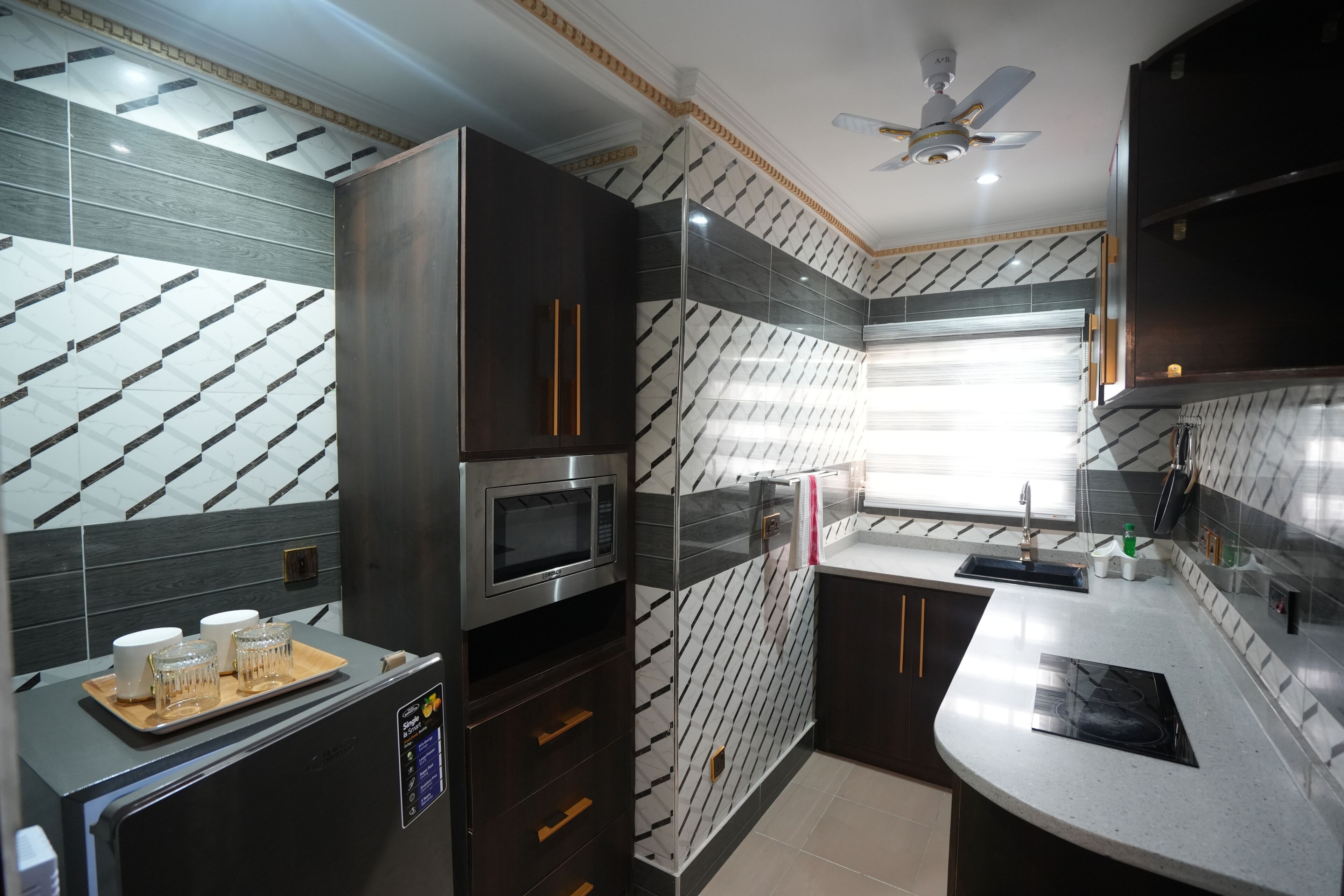 Una cocina moderna con un microondas Bosch, armarios oscuros, una ventana con persianas y un ventilador de techo.