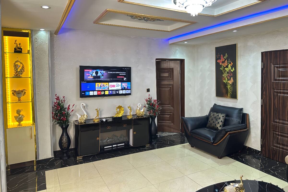 Uma sala de estar moderna com um sofá de couro preto, uma TV fixada na parede, uma mesa de console decorativa e um candelabro.
