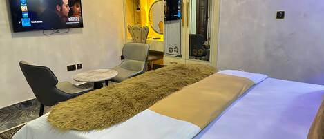 A hotel room with a bed, a TV mounted on the wall, a seating area with a chair and a small table, and a mirror with decorative elements.