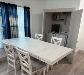 Un coin repas avec une table de couleur claire et des chaises blanches, une petite cuisine équipée avec un lave-linge et un téléviseur, et une fenêtre avec des rideaux bleus.