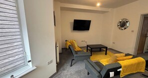 A living room with a sofa, a small table, a TV mounted on the wall, and a decorative mirror. - 1BR Parkside Apt Contractors & Families w/Parking (West Northamptonshire)