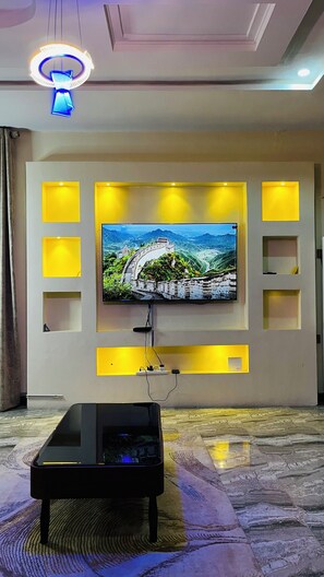 A living room with a flat-screen TV mounted on a wall with built-in shelves, a coffee table in front, and a patterned carpet. - Prime Ville
Beautiful 2-bedroom offering comfort, convenience, and peaceful stay (Lugbe)