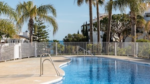 A swimming pool with a metal ladder, surrounded by a white fence, palm trees, and buildings in the background. - AS EXT Calahonda MAR 10 (Mijas)