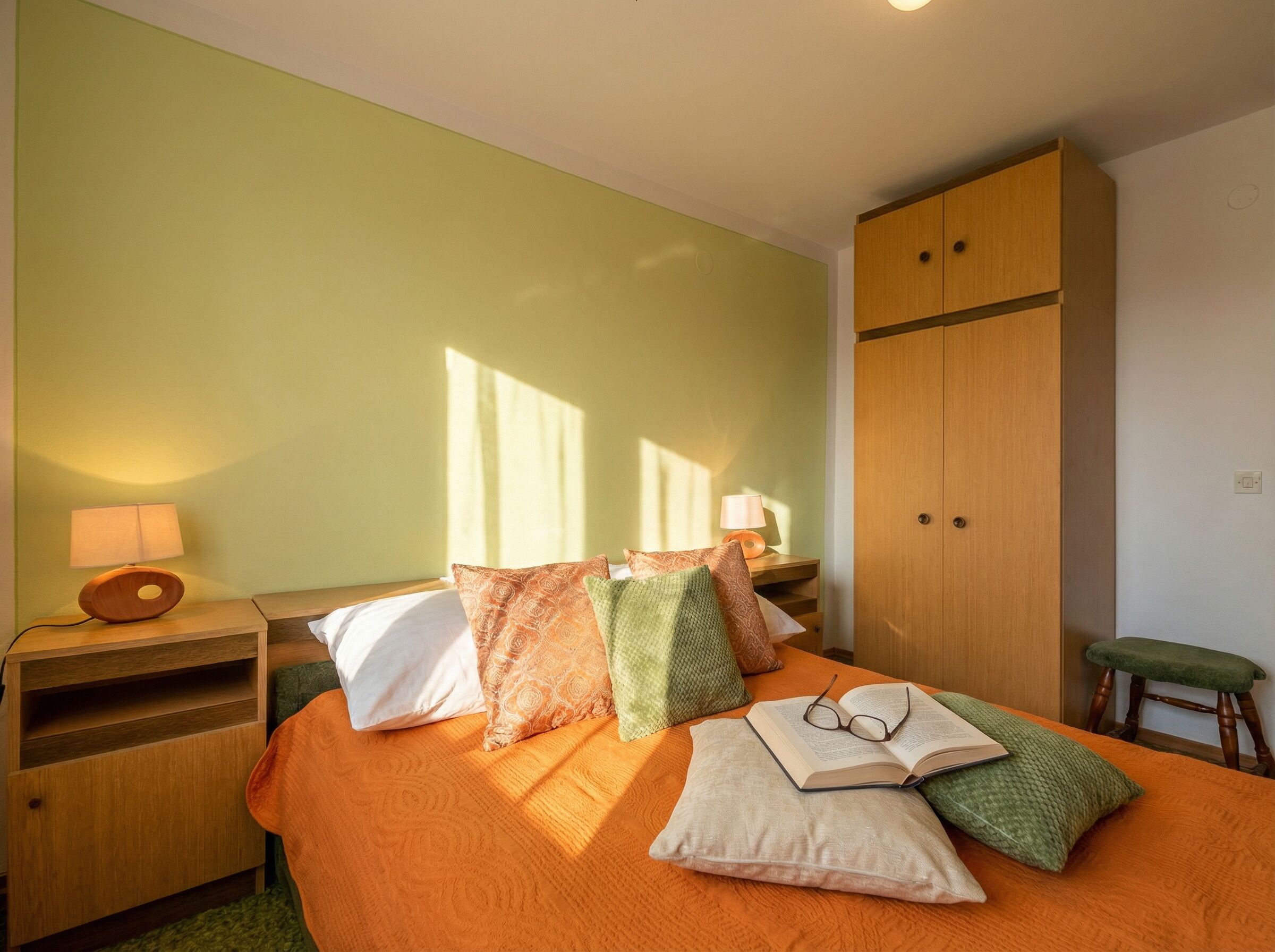 Ein Schlafzimmer mit einem Bett, Kissen, einem Nachttisch mit Lampe, einem Holzschrank und einem grünen Sessel.