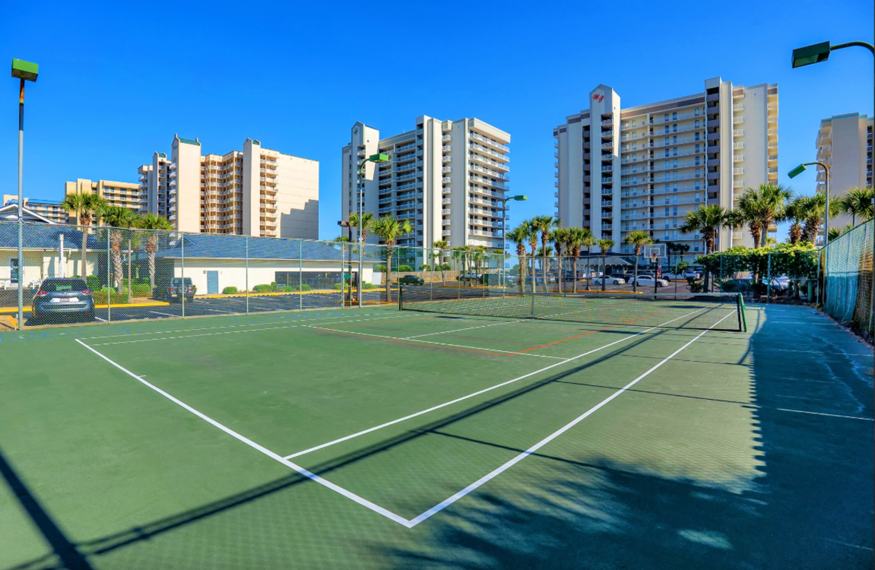 An outdoor tennis court with a net, surrounded by a fence and palm trees, with buildings in the background.