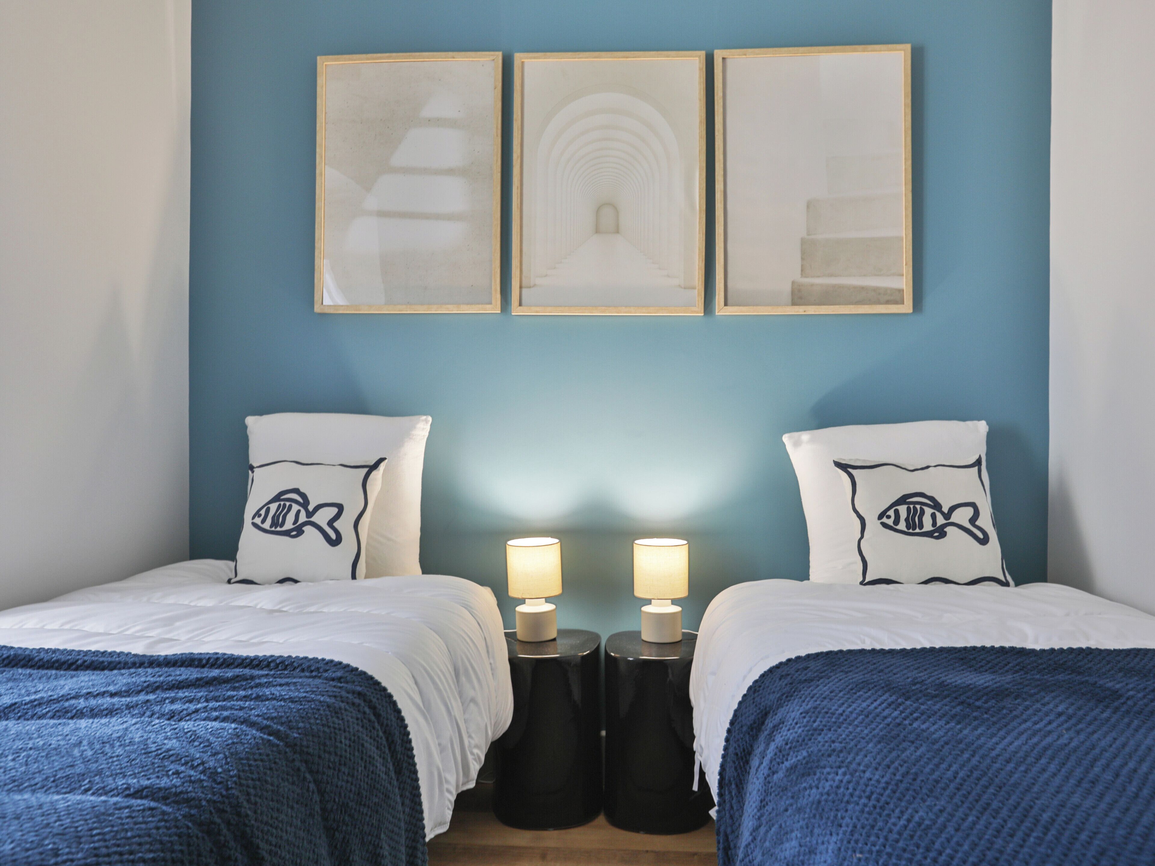 Une chambre d’hôtel avec deux lits, un mur bleu et trois tableaux encadrés au-dessus des lits.