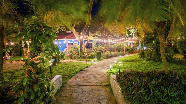 Un sendero de piedra conduce a un edificio con tejado rojo, rodeado de vegetación exuberante y luces decorativas.