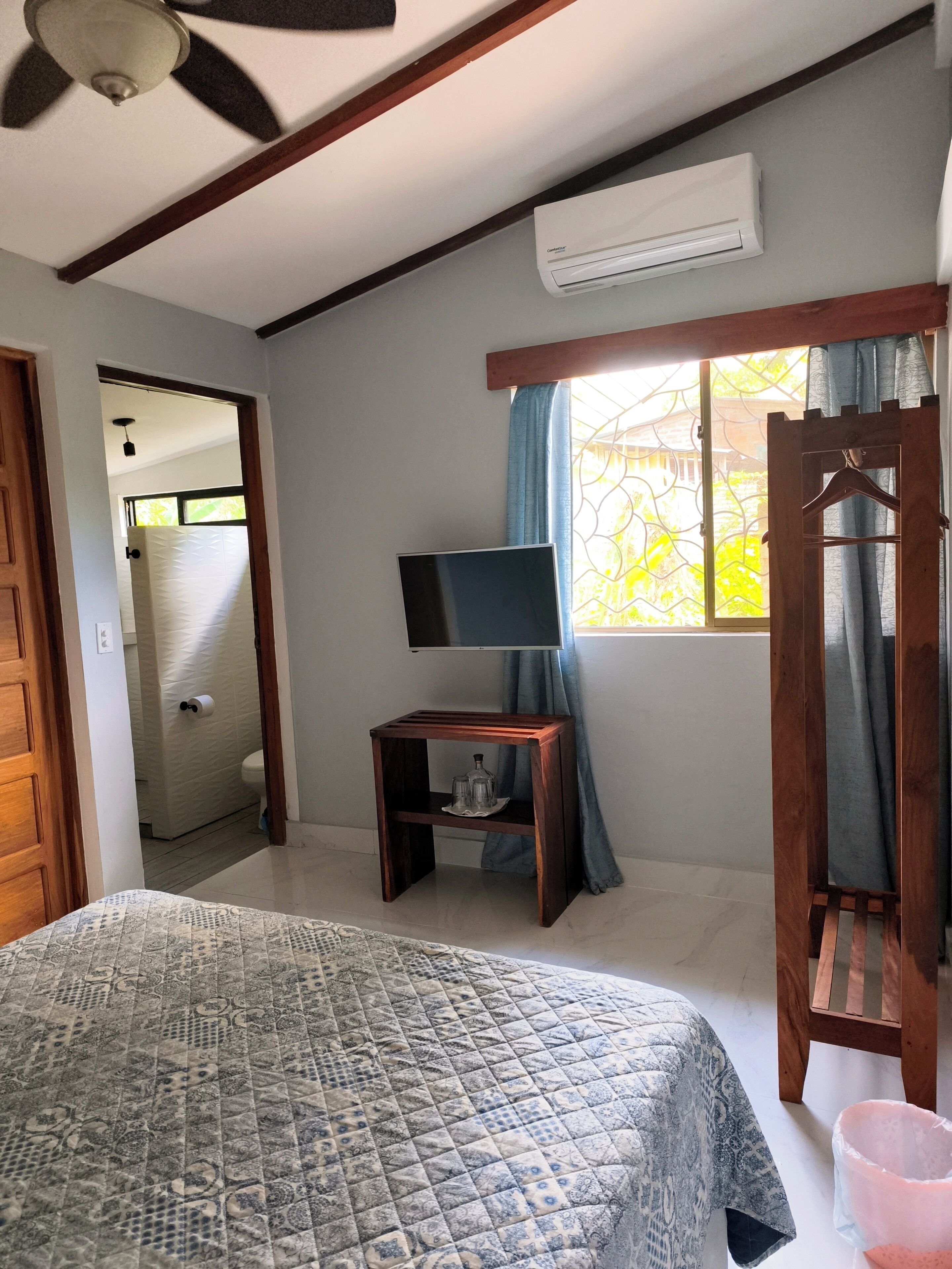 A bedroom with a bed, a TV on a stand, an air conditioner, a window with curtains, and a wooden door.