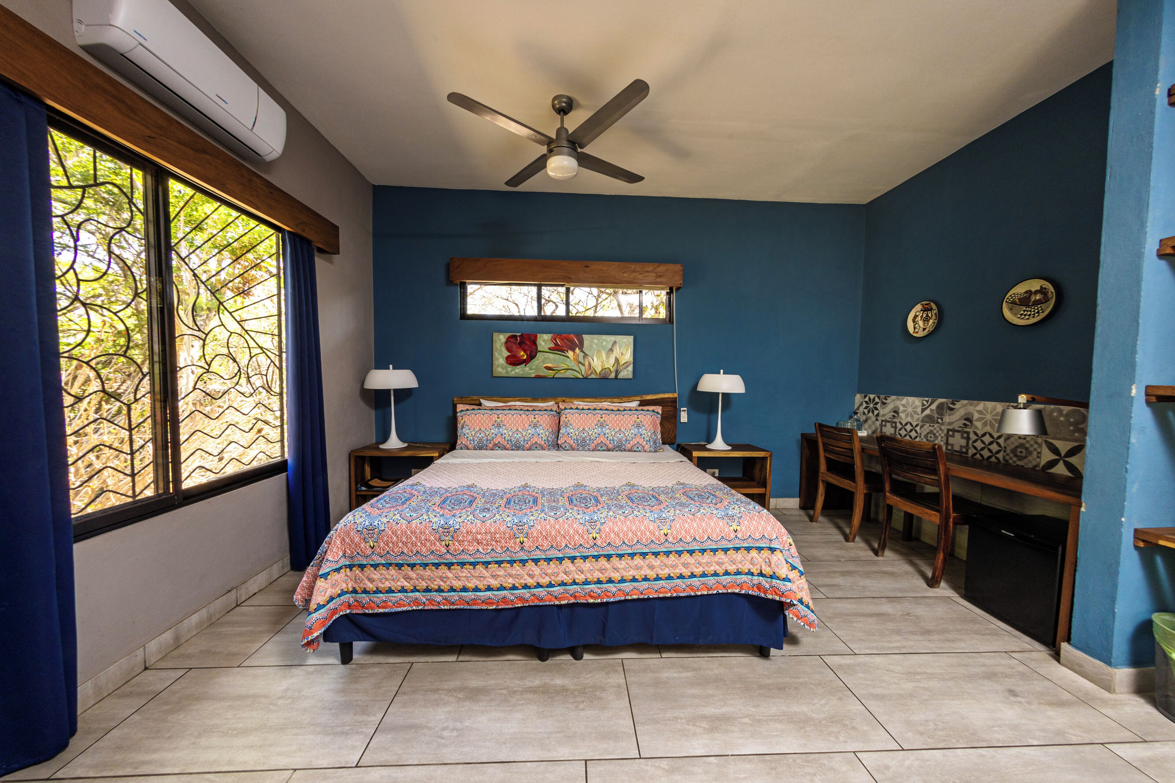 A bedroom with a bed, two bedside tables, a ceiling fan, a window with curtains, a painting, a desk with a chair, and a tiled floor.