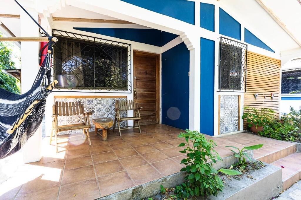Una casa con porche, una hamaca, dos sillas de madera y piso de baldosas.