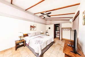 Une chambre à coucher comprenant un lit, des tables de chevet, un ventilateur de plafond, une télévision et une porte donnant sur une autre pièce.