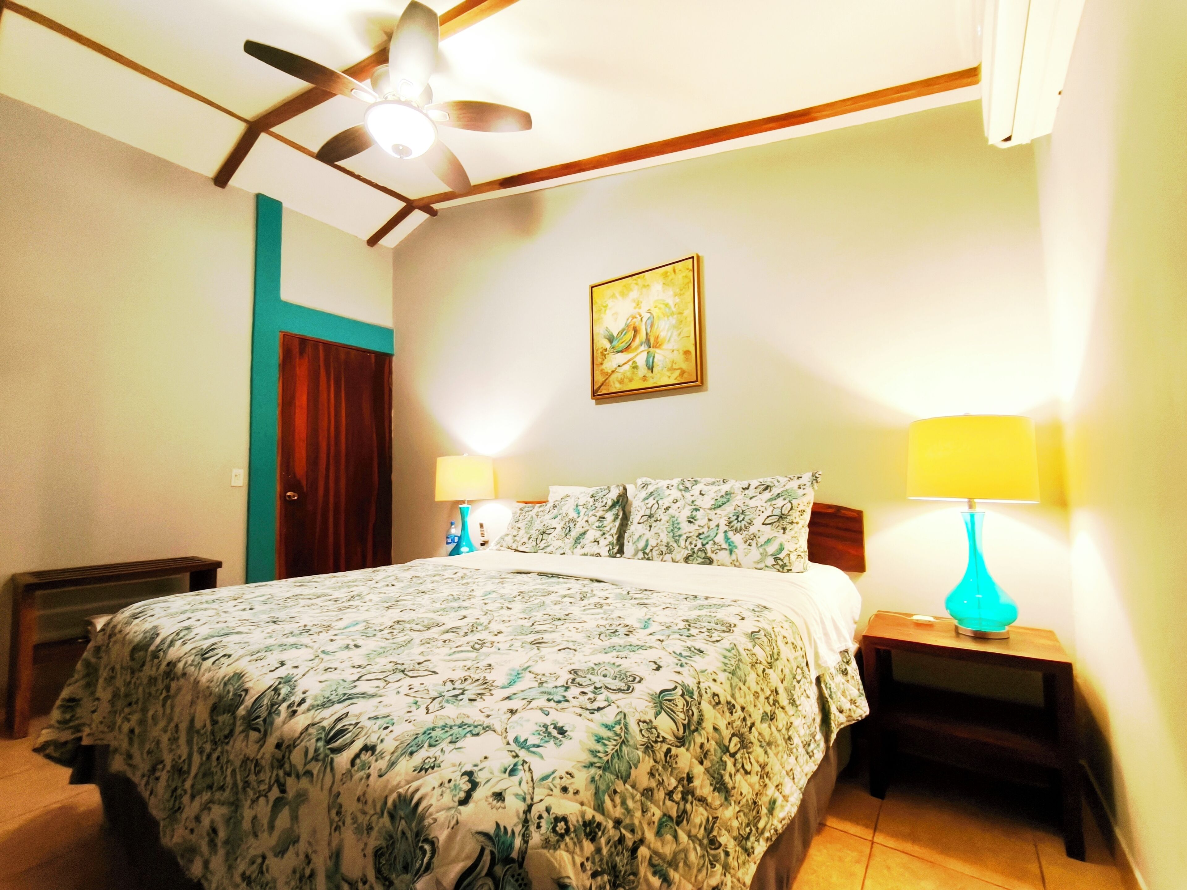A bedroom with a bed, two bedside tables with lamps, a ceiling fan, a door, and a painting on the wall.