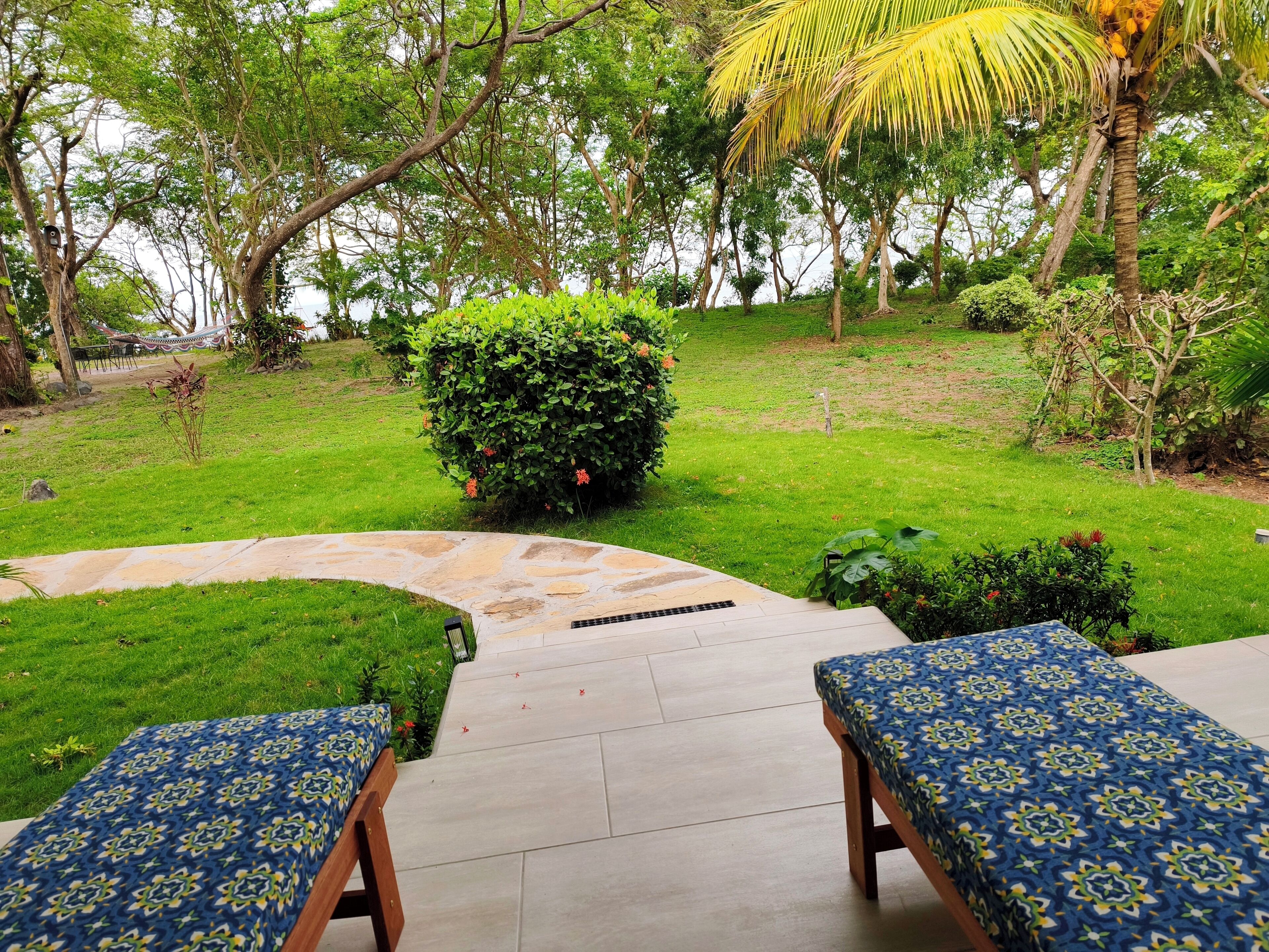 A patio area with stone flooring, two wooden benches with patterned cushions, and a lush green garden with various plants and a palm tree.