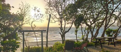 A stone-paved patio with a wooden pergola, a bench, and a hammock overlooking a beach.