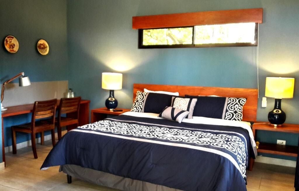 Un dormitorio con una cama, dos mesitas de noche con lámparas, un escritorio con silla y una estantería en la pared.