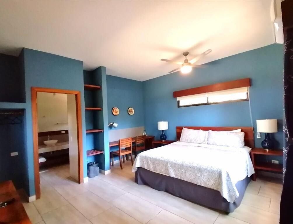 Une chambre à coucher comprenant un lit, des tables de chevet, un bureau, une chaise, un ventilateur de plafond, et une salle de bain visible à travers une porte ouverte.