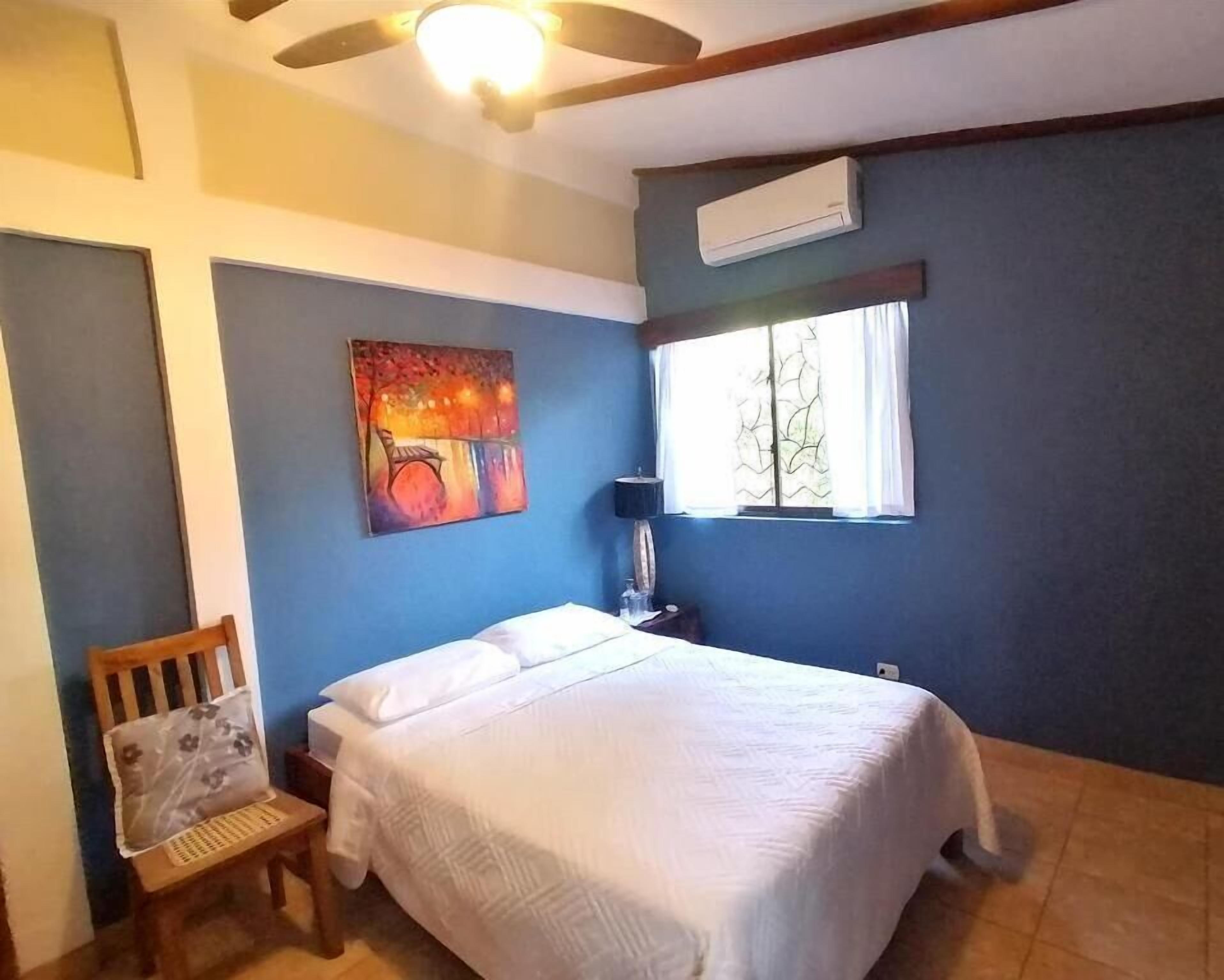 Een slaapkamer met een bed, een stoel, een schilderij, een raam en een airconditioning.