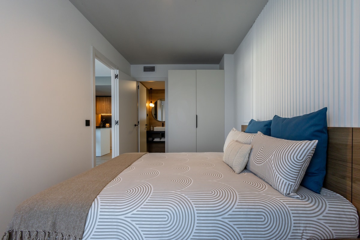 A bedroom with a bed, pillows, a bedside table, and a view into the bathroom.