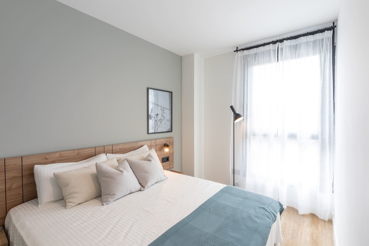 A bedroom with a bed, pillows, a bedside lamp, a framed picture on the wall, and a window with curtains.