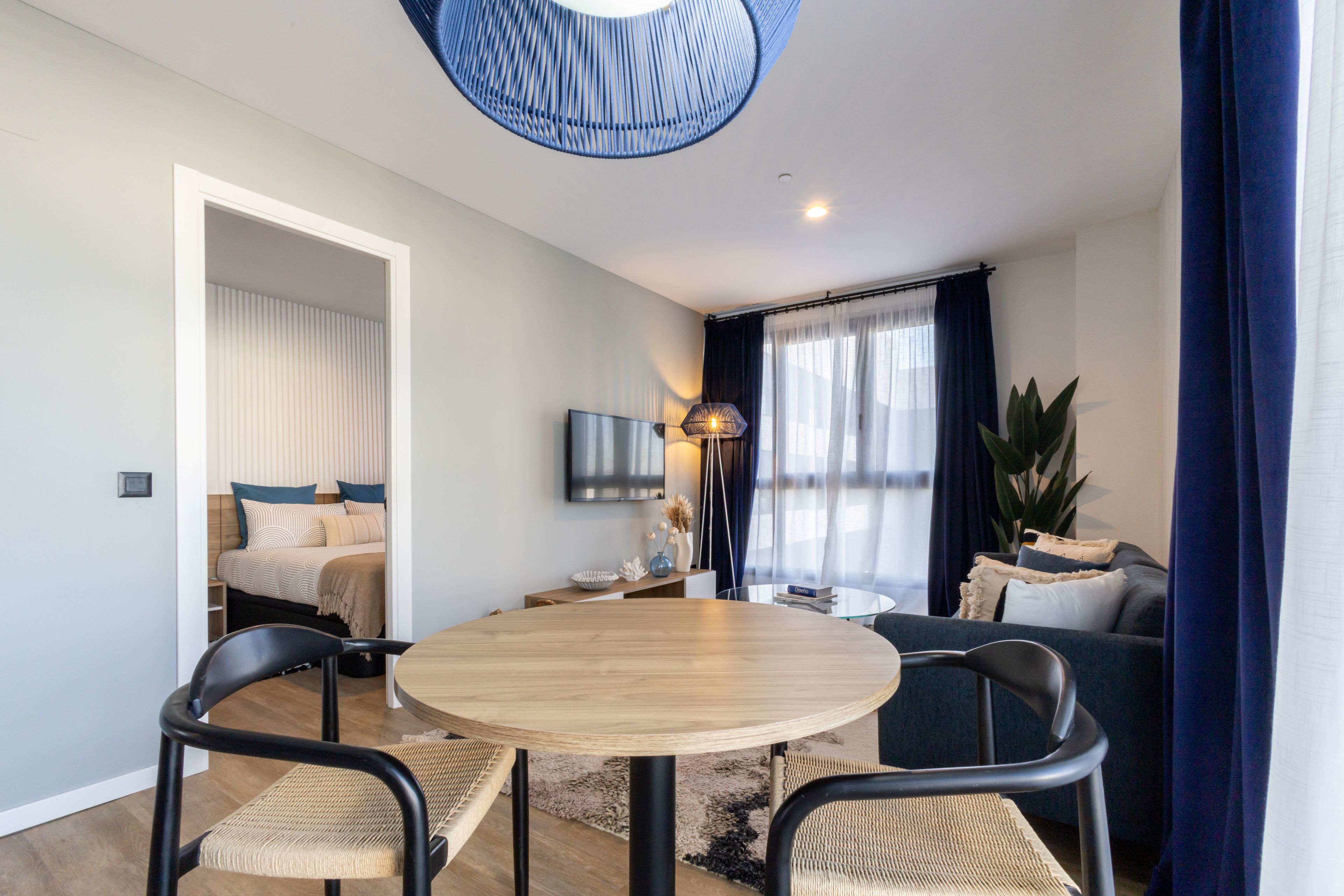 Een moderne woonkamer met een ronde houten tafel, twee stoelen, een bank, een aan de muur bevestigde televisie en een groot raam met gordijnen.